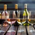 best wine to pair with BBQ - Six wine glasses filled with different wines—Zinfandel, Rosé, Chardonnay, Sparkling Wine, Riesling, and Cabernet Sauvignon—on a wooden BBQ table with matching wine bottles in the background.